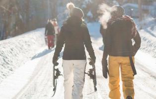 Im Vordergrund befinden zwei Menschen in Winterkleidung, von hinten fotografiert, die über einen verschneiten, sonnigen Waldweg laufen. Die Person links hält Schneeschuhe in den Händen. Im Hintergrund sind drei weitere Menschen zu sehen. 